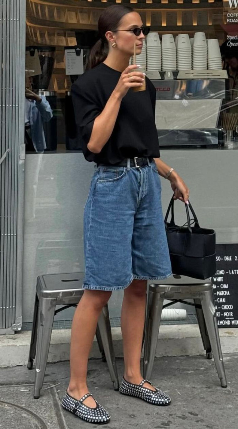 Woman in black top and blue denim shorts standing outdoors, holding a drink.
